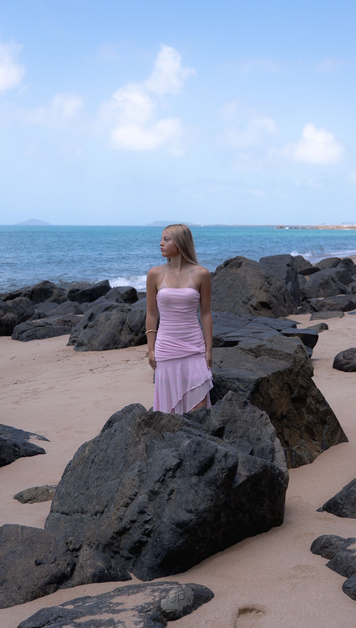 A figure in a pink dress sits thoughtfully on dark volcanic rocks along a sandy beach with turquoise ocean waters.