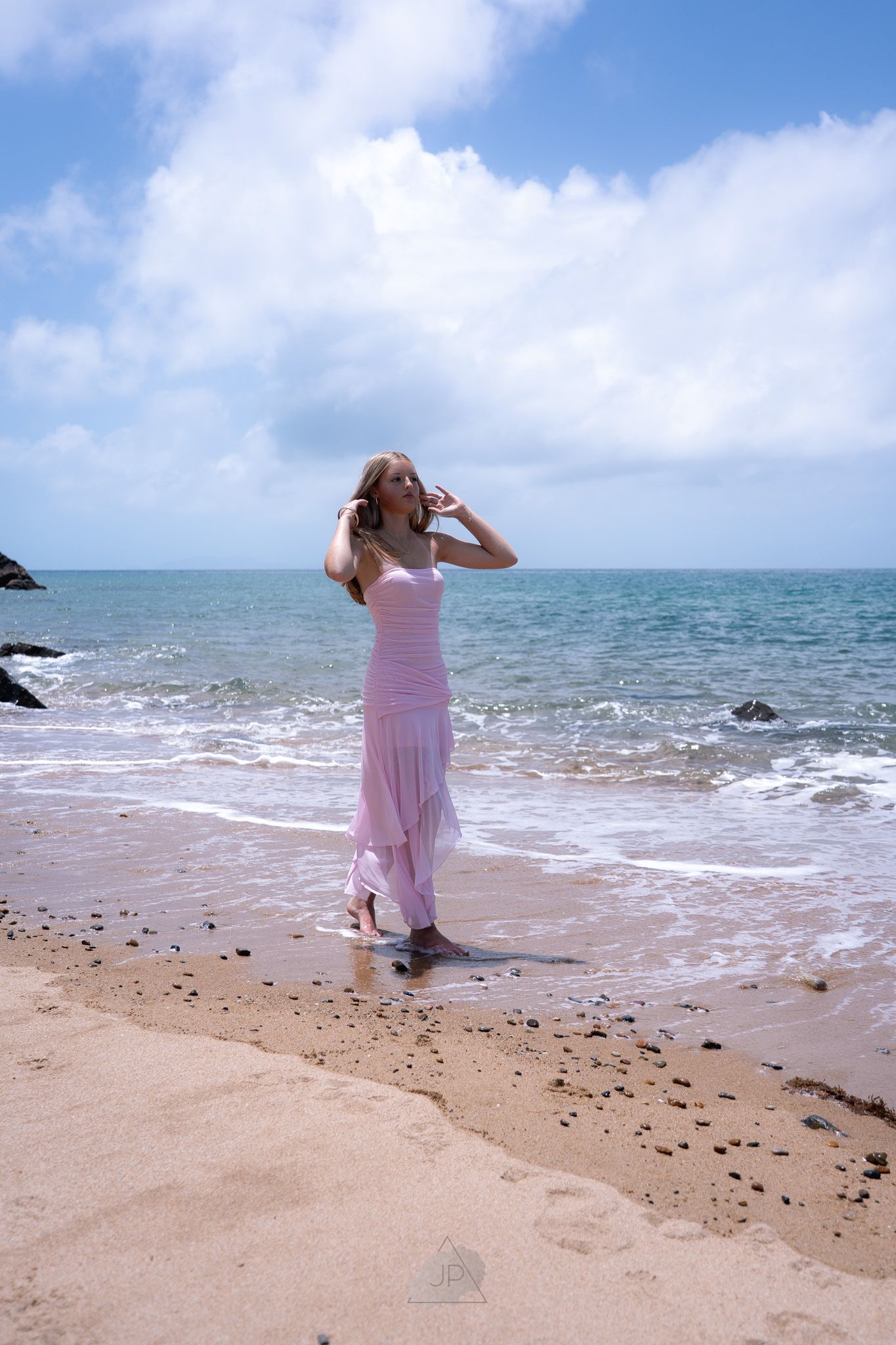 Person in pink dress stands on rocky beach taking photos of ocean waves under cloudy sky.
