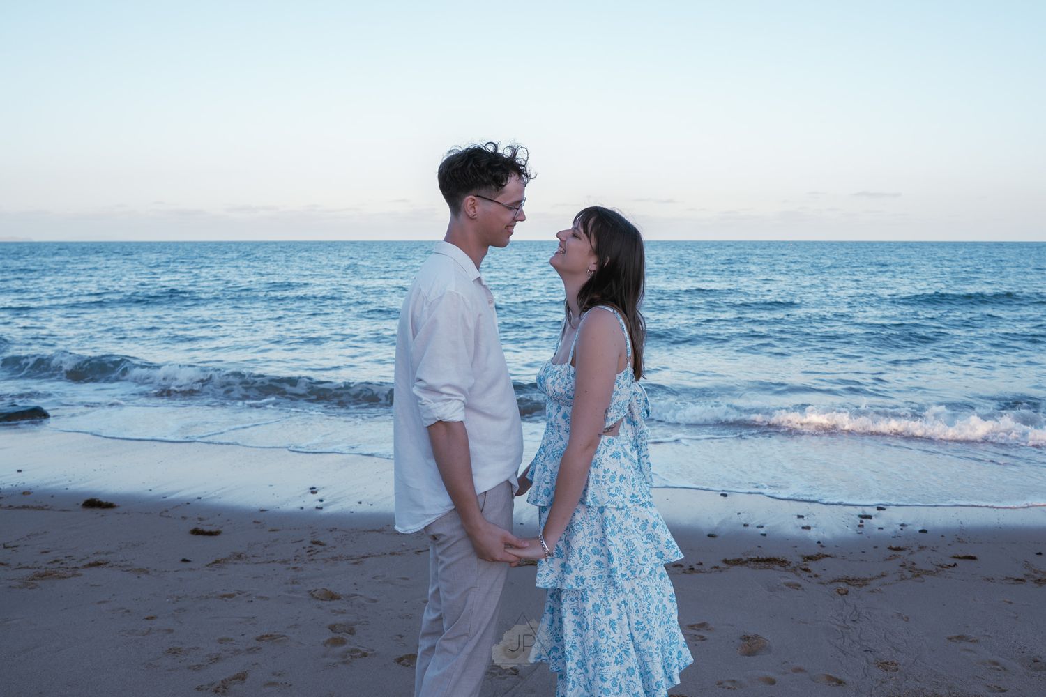 A couple stands together on a beach at sunset holding hands against the blue ocean backdrop.