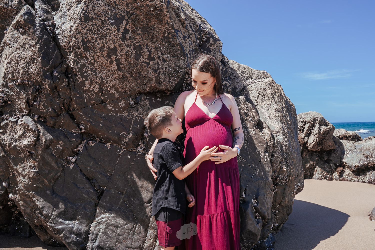 Two figures in a pink dress and black outfit stand together against large coastal rocks with ocean in the background.