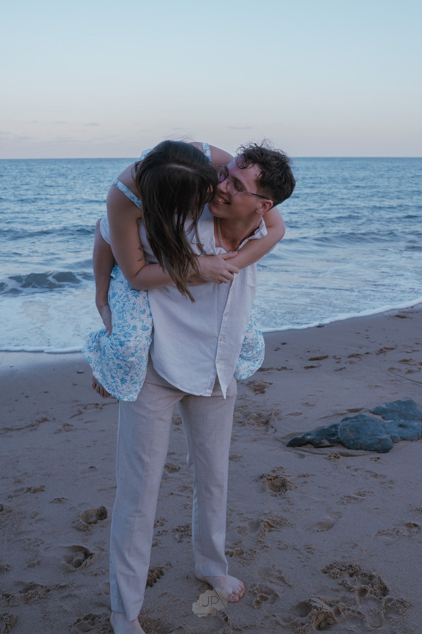 A romantic couple shares a tender dip kiss on a sandy beach at sunset with ocean waves in the background.