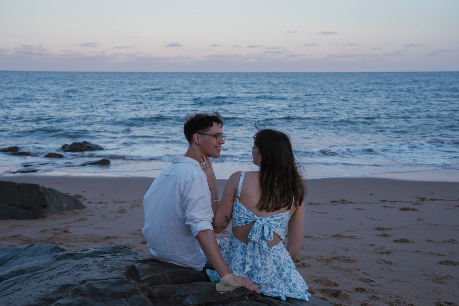 A couple sits together on a beach at sunset with waves crashing on the rocky shore in the background.