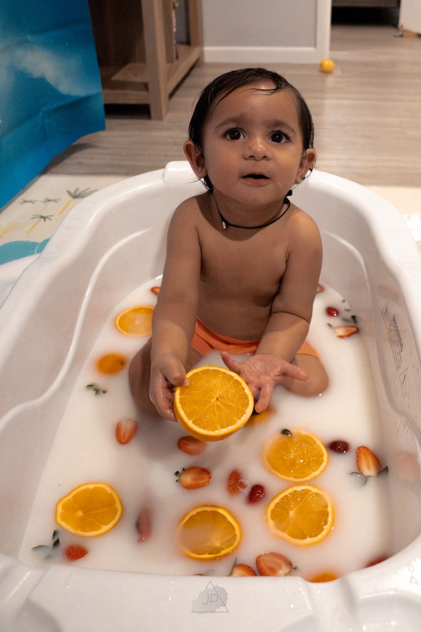 A small child sits in a white bathtub with orange slices and rubber ducks for a creative fruit-themed bath photoshoot.