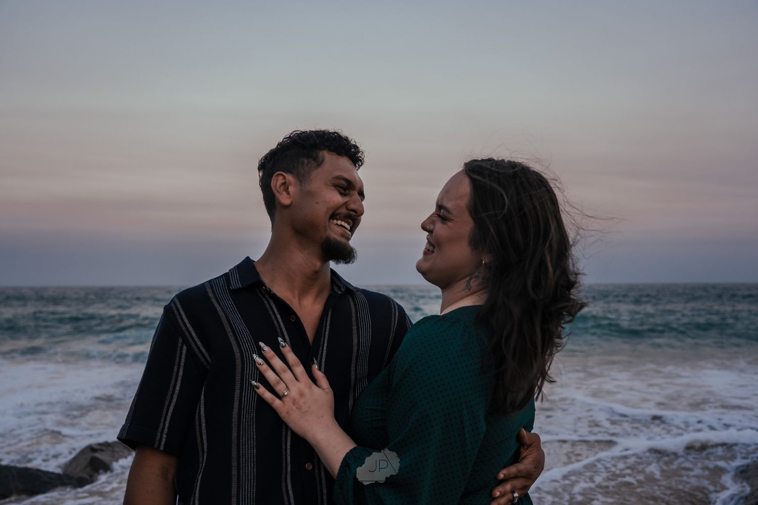 Couple sharing intimate moments at sunset along a rocky beach coastline with crashing waves.