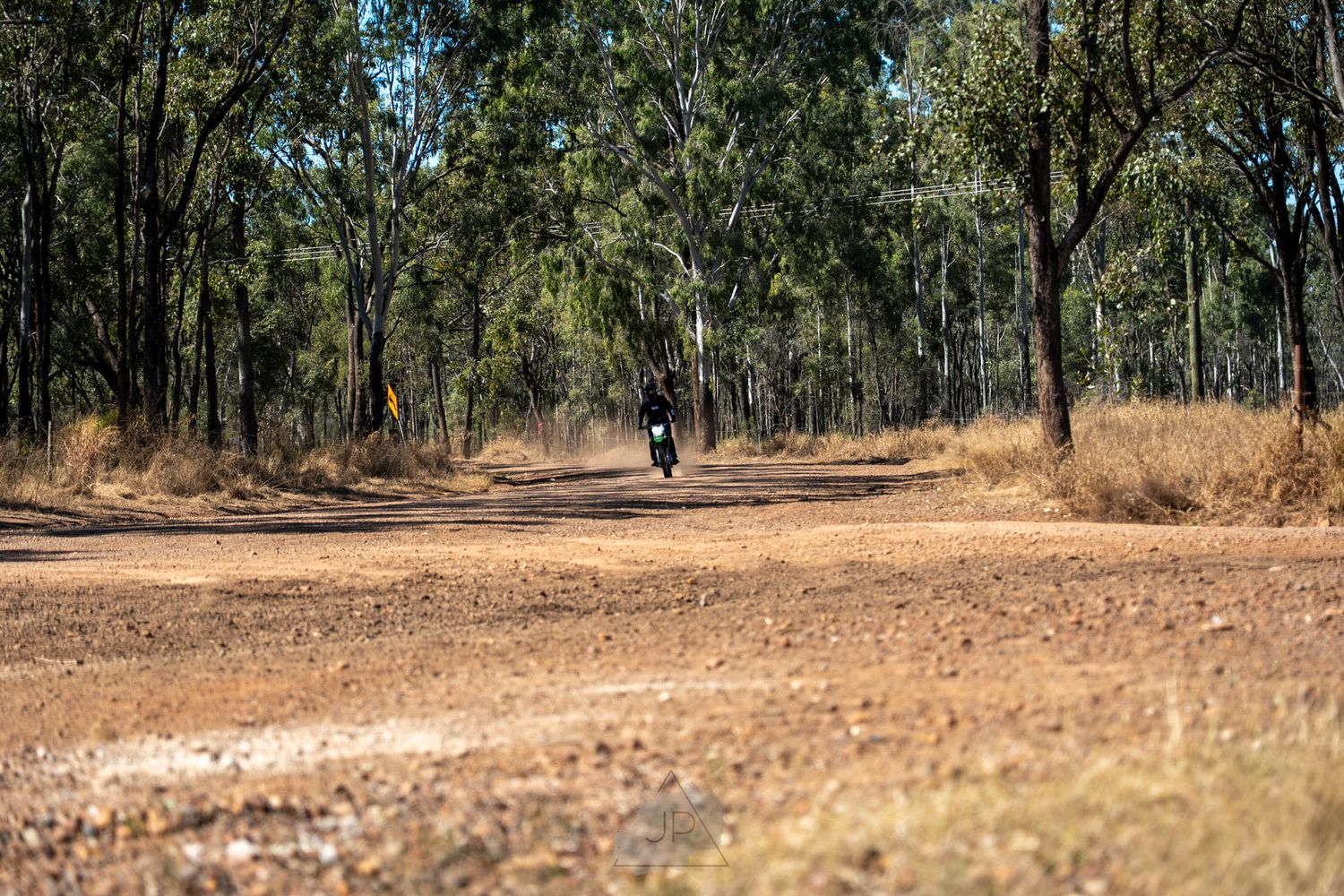 A scenic country dirt road winding through dry eucalyptus woodland under a bright blue Australian sky.