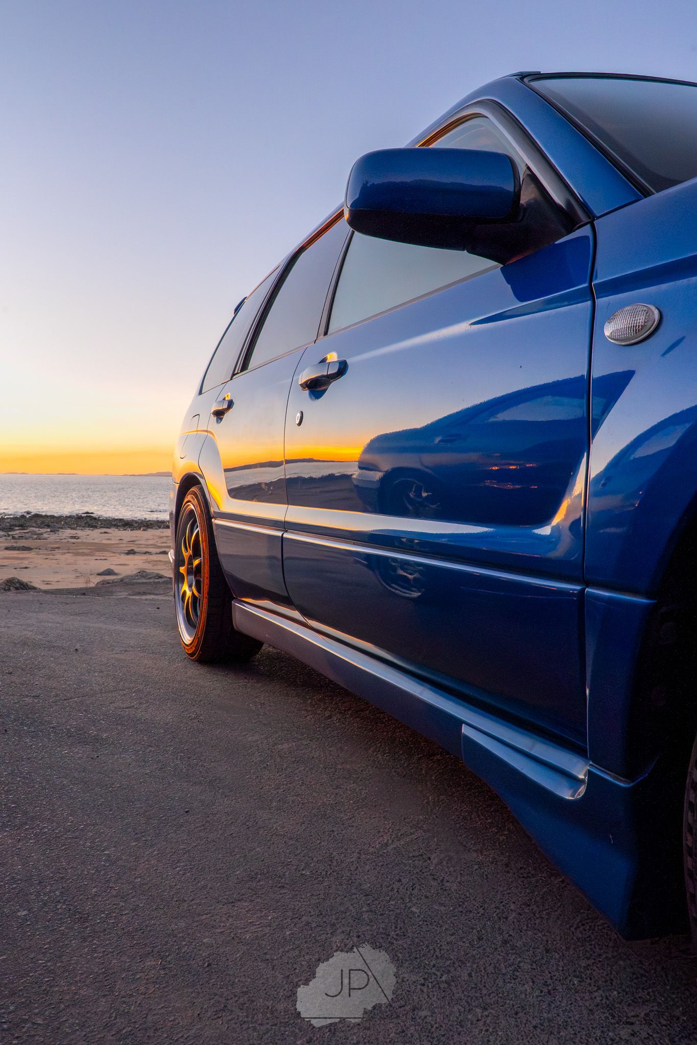 Blue sports car photographed at low angle against desert sunset showing sleek side profile and custom wheels.
