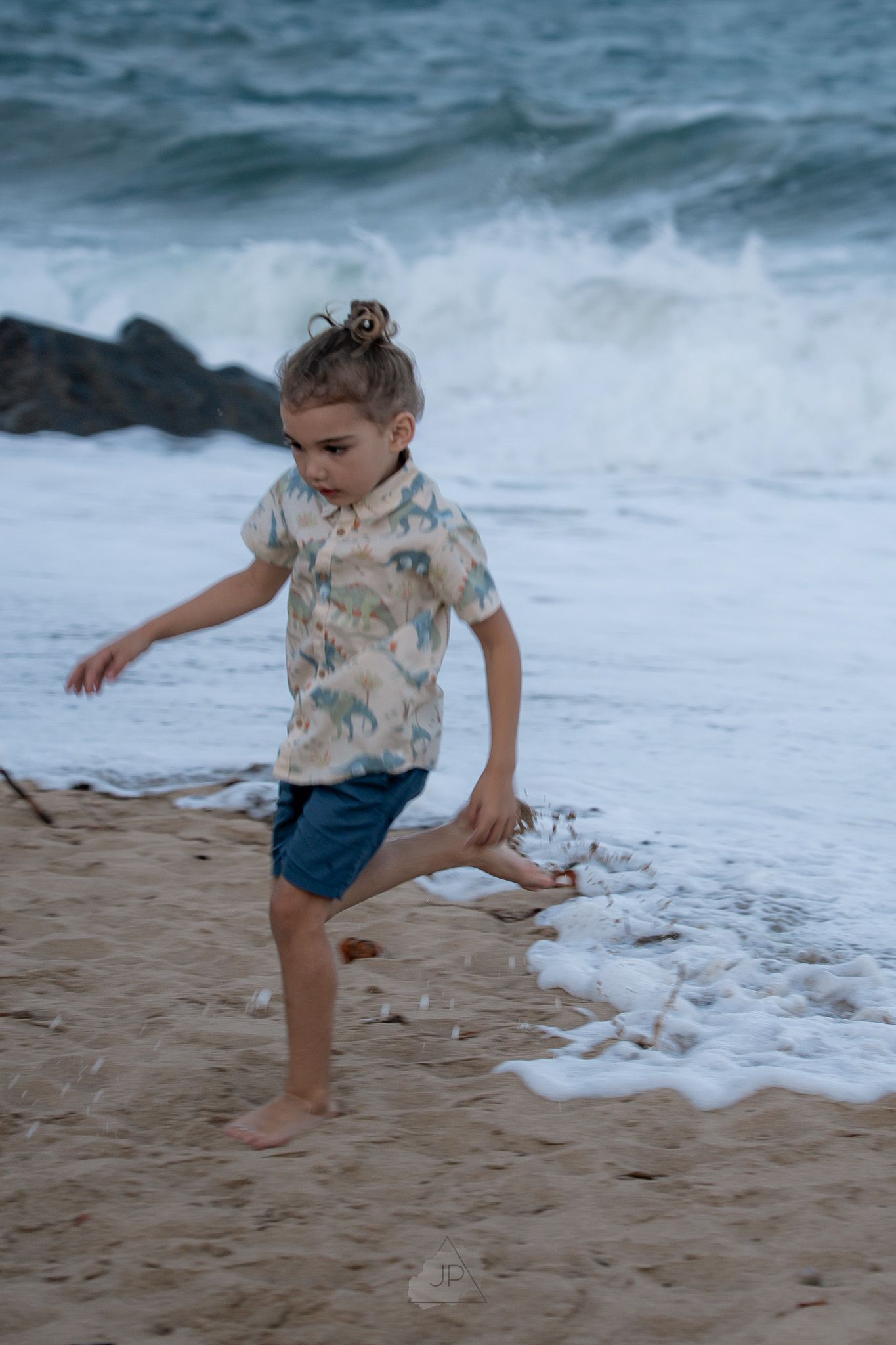 A child in a floral shirt and blue shorts plays along the ocean shoreline, running from incoming waves on the beach.
