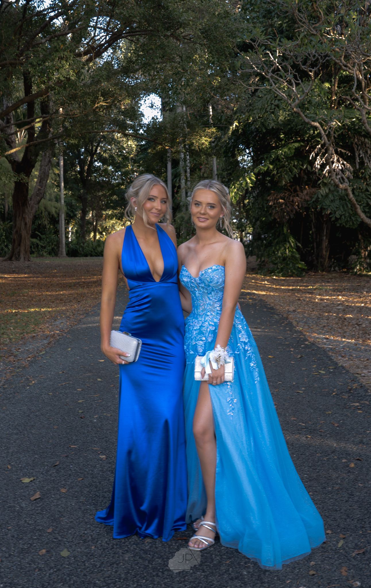 Two formal dresses in shades of blue - a royal blue fitted gown and a light blue sparkly gown with a slit on a forest path.