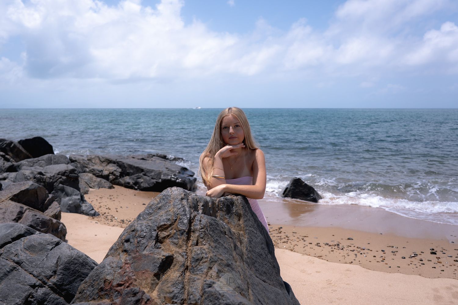 A person sitting on rocky coastal cliffs overlooking the turquoise ocean waves on a sunny day.