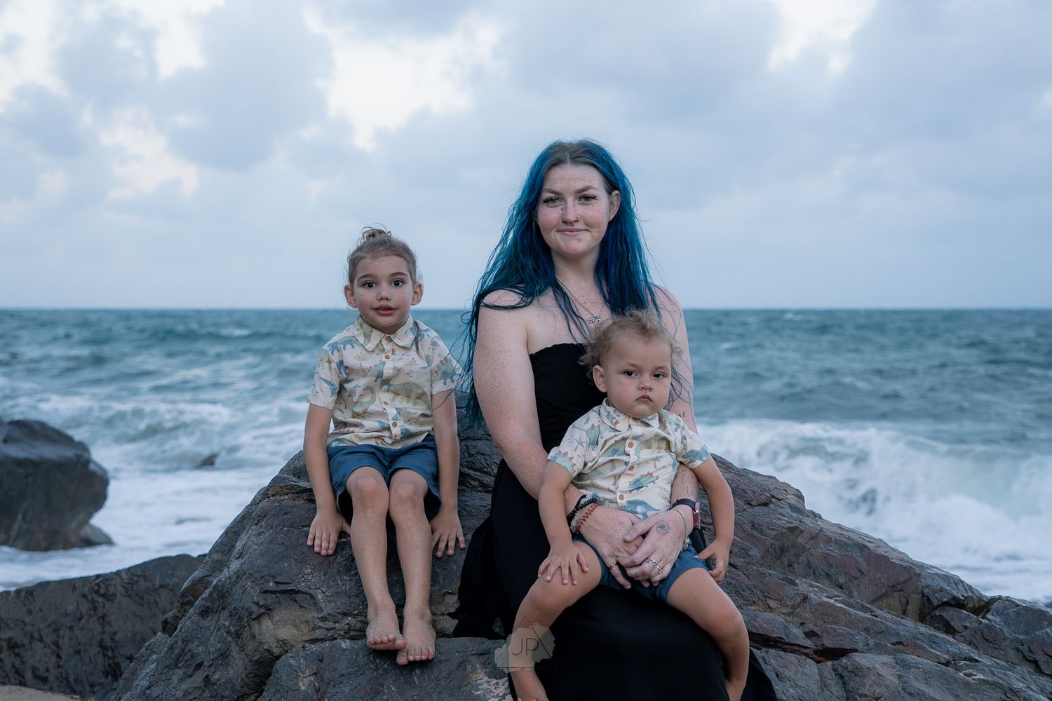A family enjoys a peaceful moment together sitting on rocks by the ocean waves at sunset.