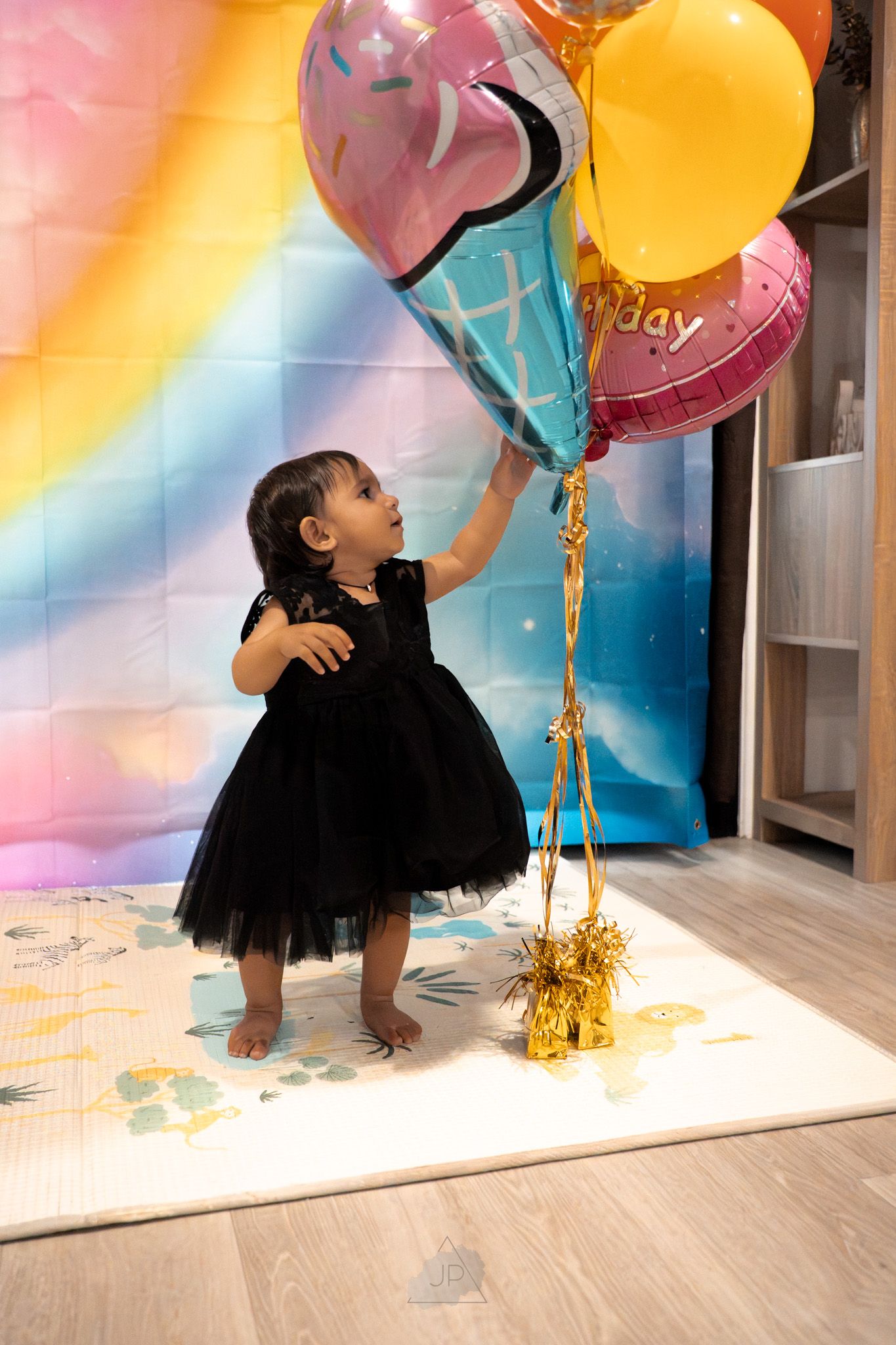 Colorful birthday photoshoot setup with balloons and a black tutu against a rainbow-colored backdrop on light wooden floors.