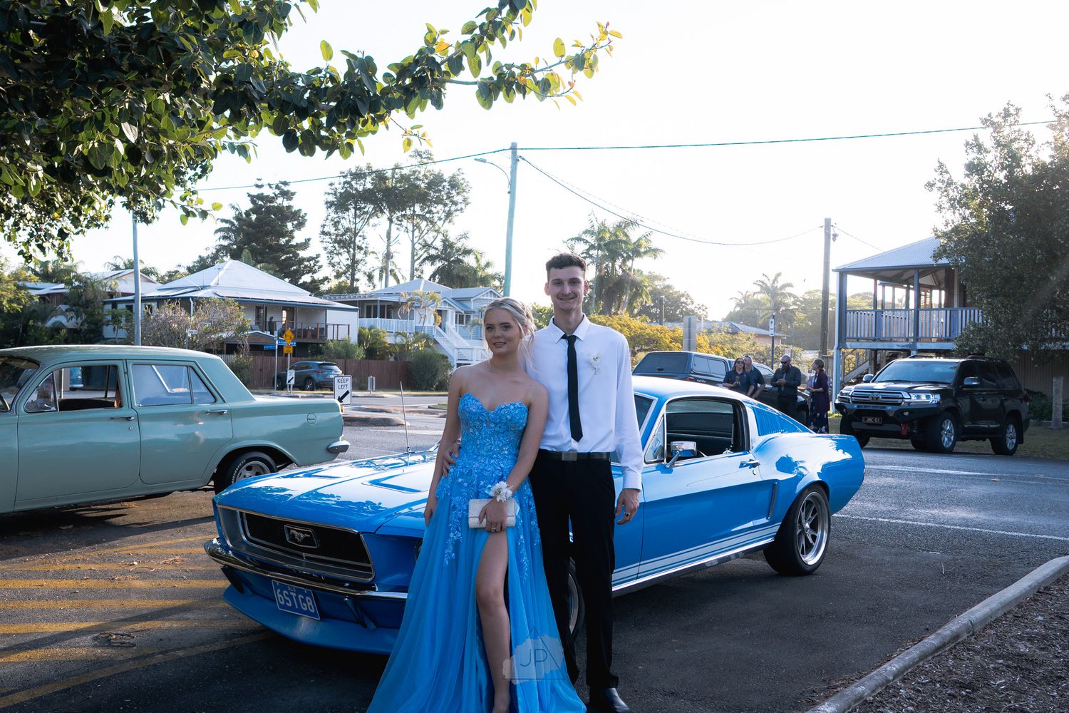 A bright blue classic Ford Mustang is displayed in a parking lot with several people gathered around it.