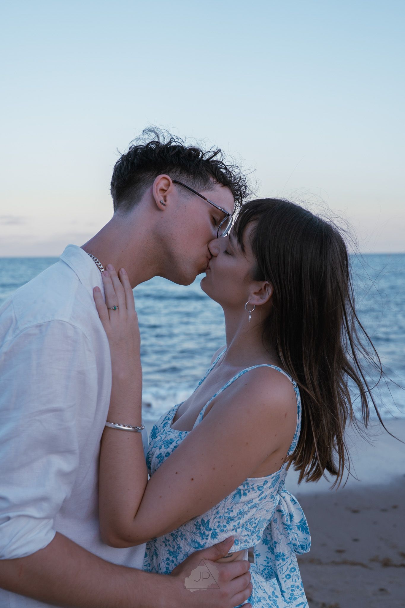 A romantic couple shares intimate kisses on a beach at sunset with ocean waves in the background.