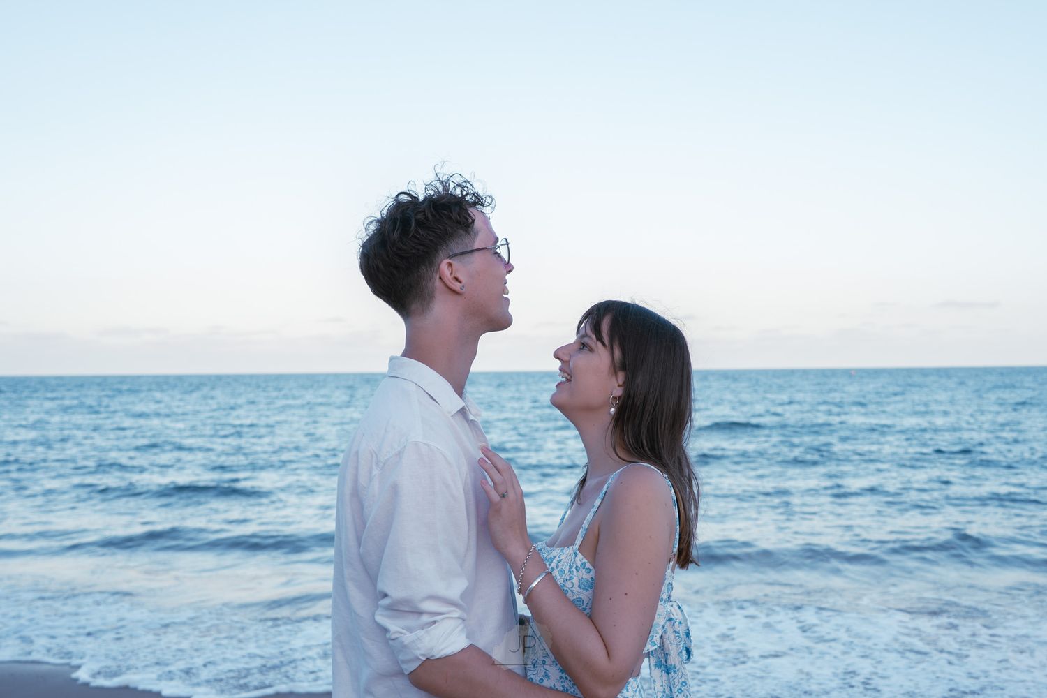 A couple embraces on the beach at sunset with ocean waves in the background.