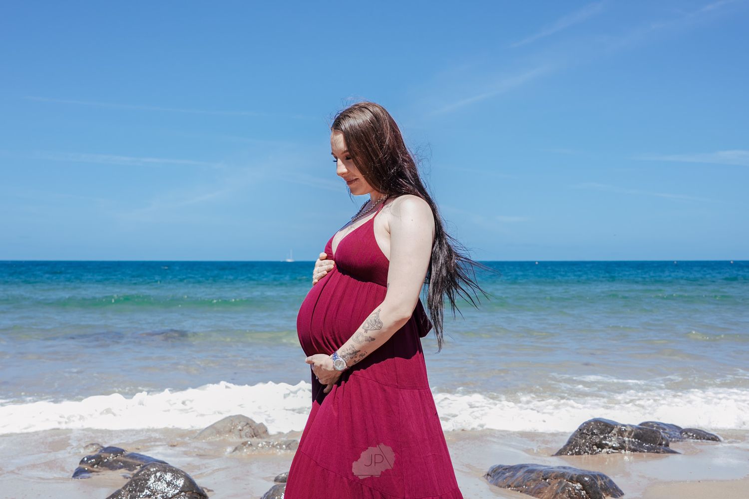 A person in a red dress poses on a scenic beach with turquoise waters and rocky outcrops in the background.
