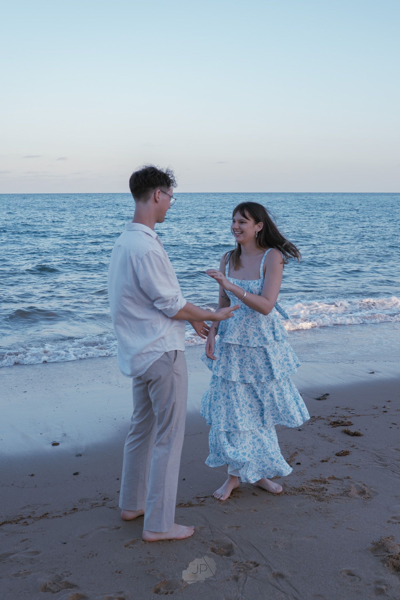 A couple in light summer attire stands together on a sandy beach with the ocean in the background.