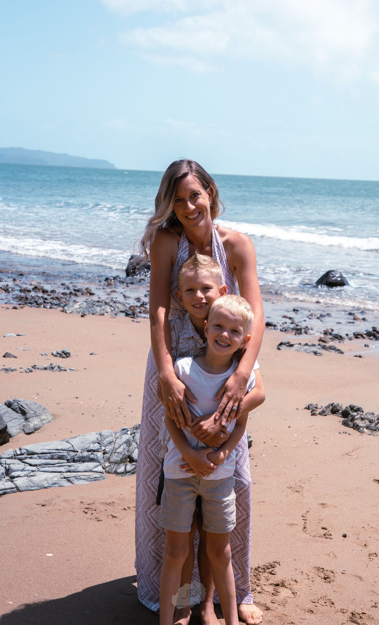Playful moment captured on a sunny beach with sand, ocean waves, and rocky shoreline in the background.