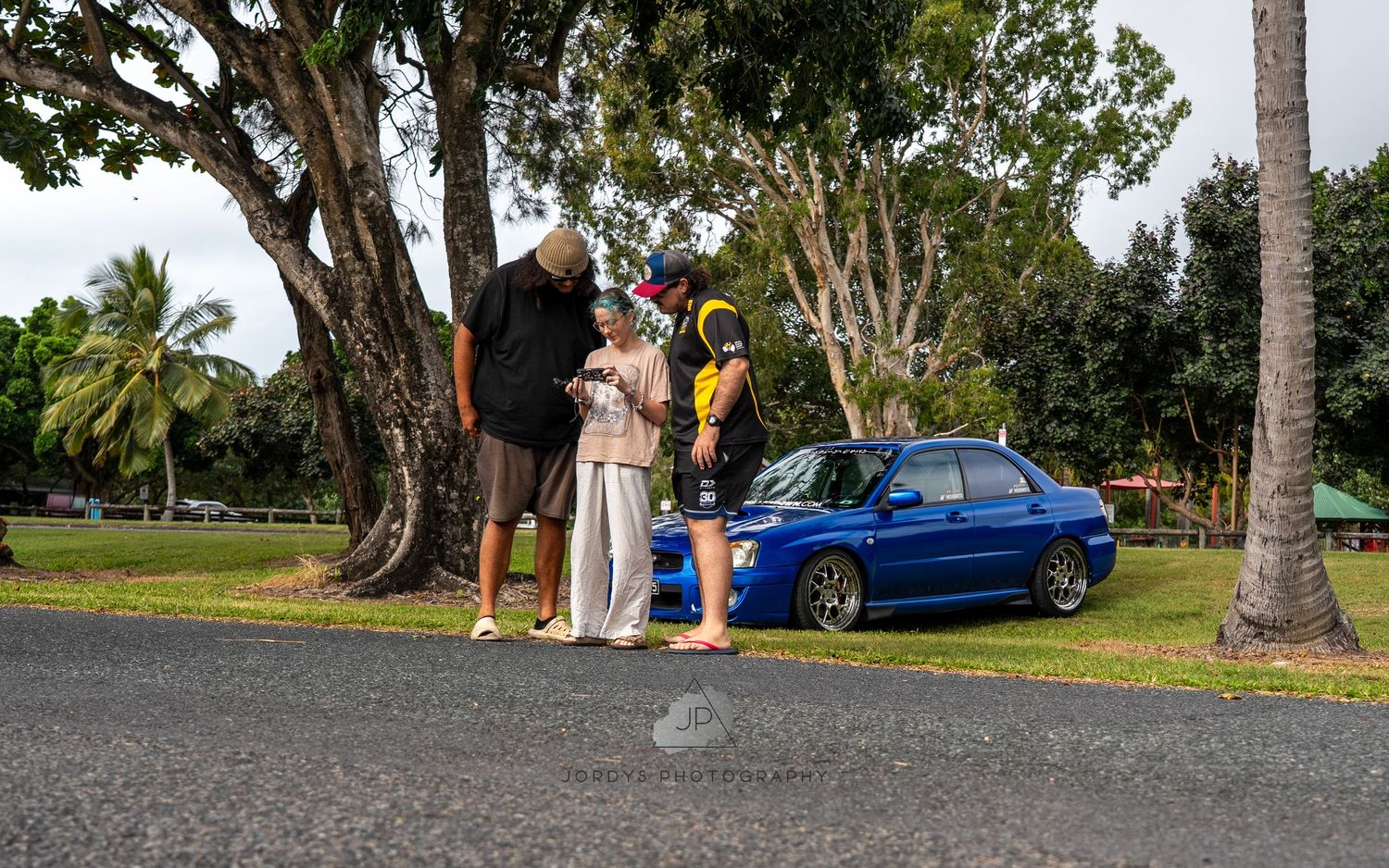 Panoramic montage of people gathering around a blue car in a lush tropical park setting with palm trees.