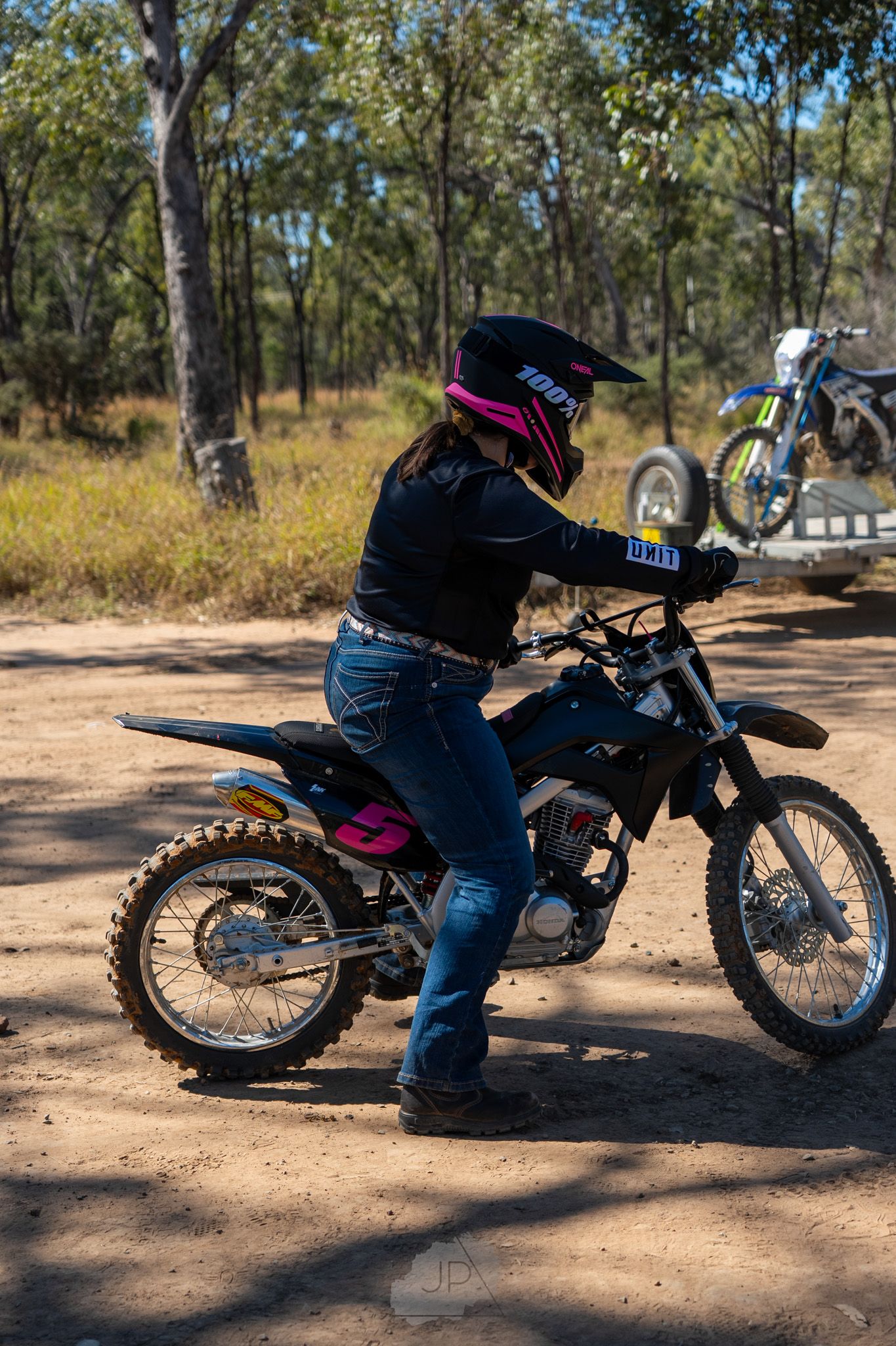 A motorcycle rider wearing protective gear sits on a dirt bike on a dusty trail surrounded by eucalyptus trees.