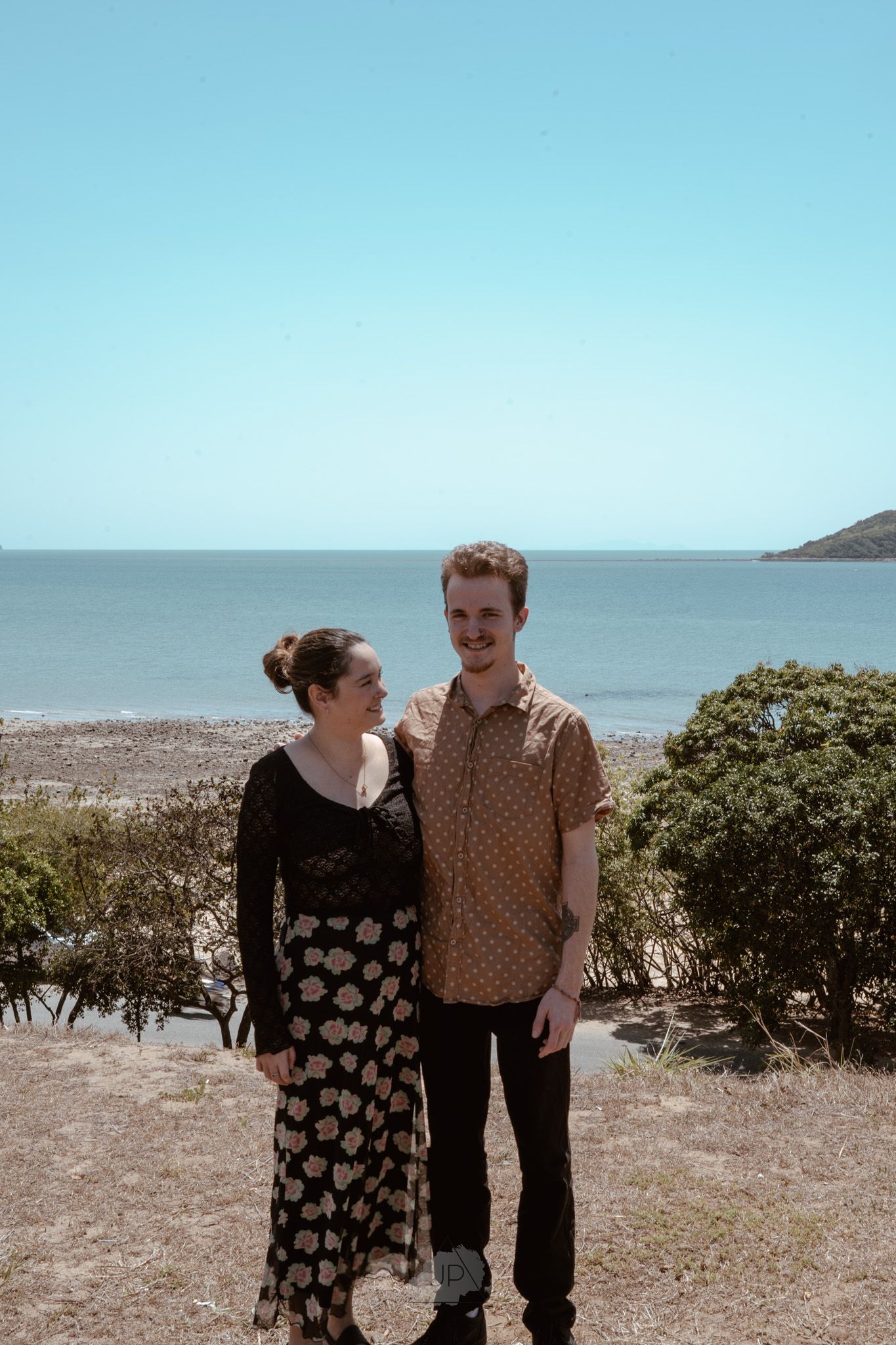 Two people stand together on a coastal hillside with the ocean and blue sky visible in the background.