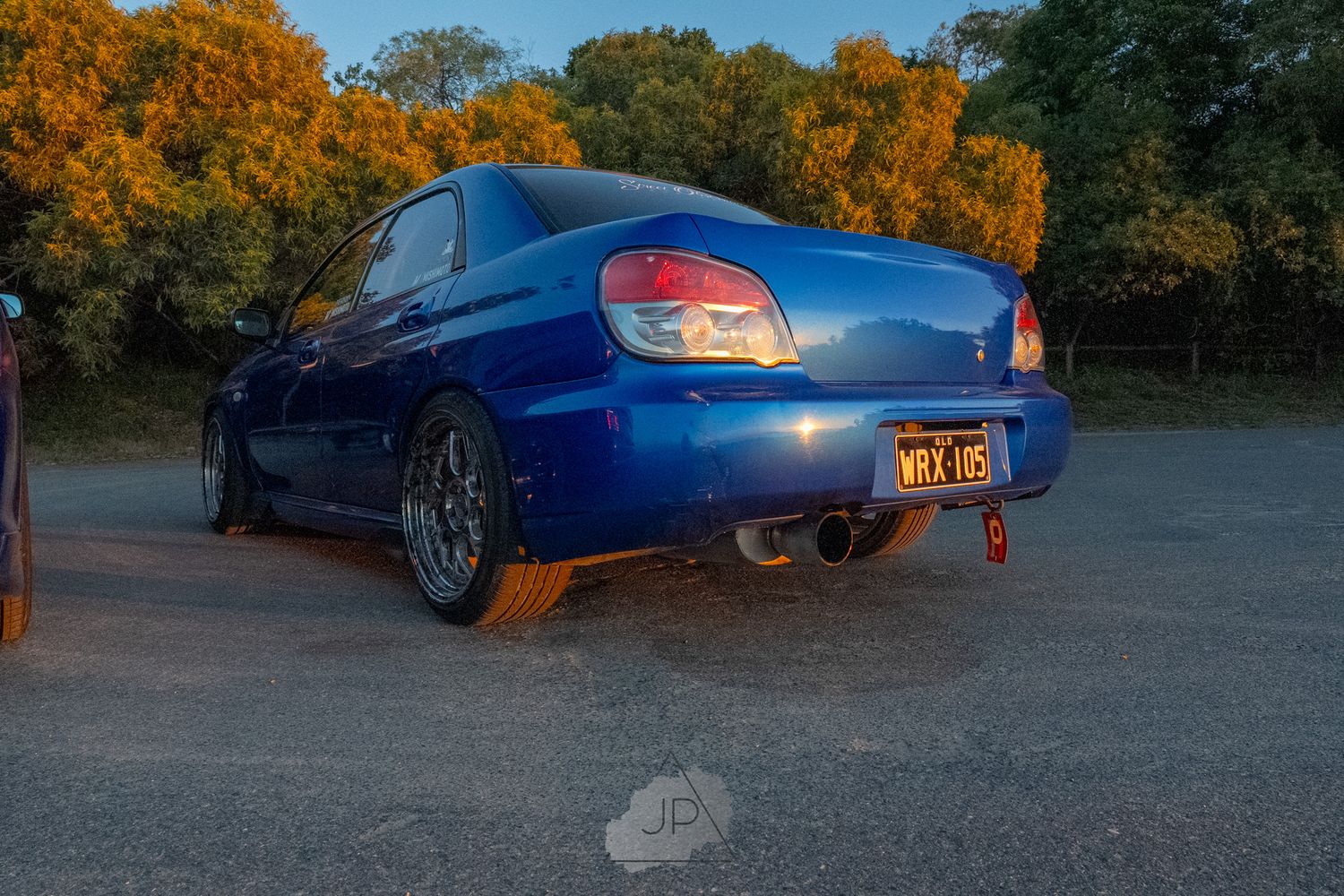 A blue sports car is parked during sunset with golden autumn trees in the background.