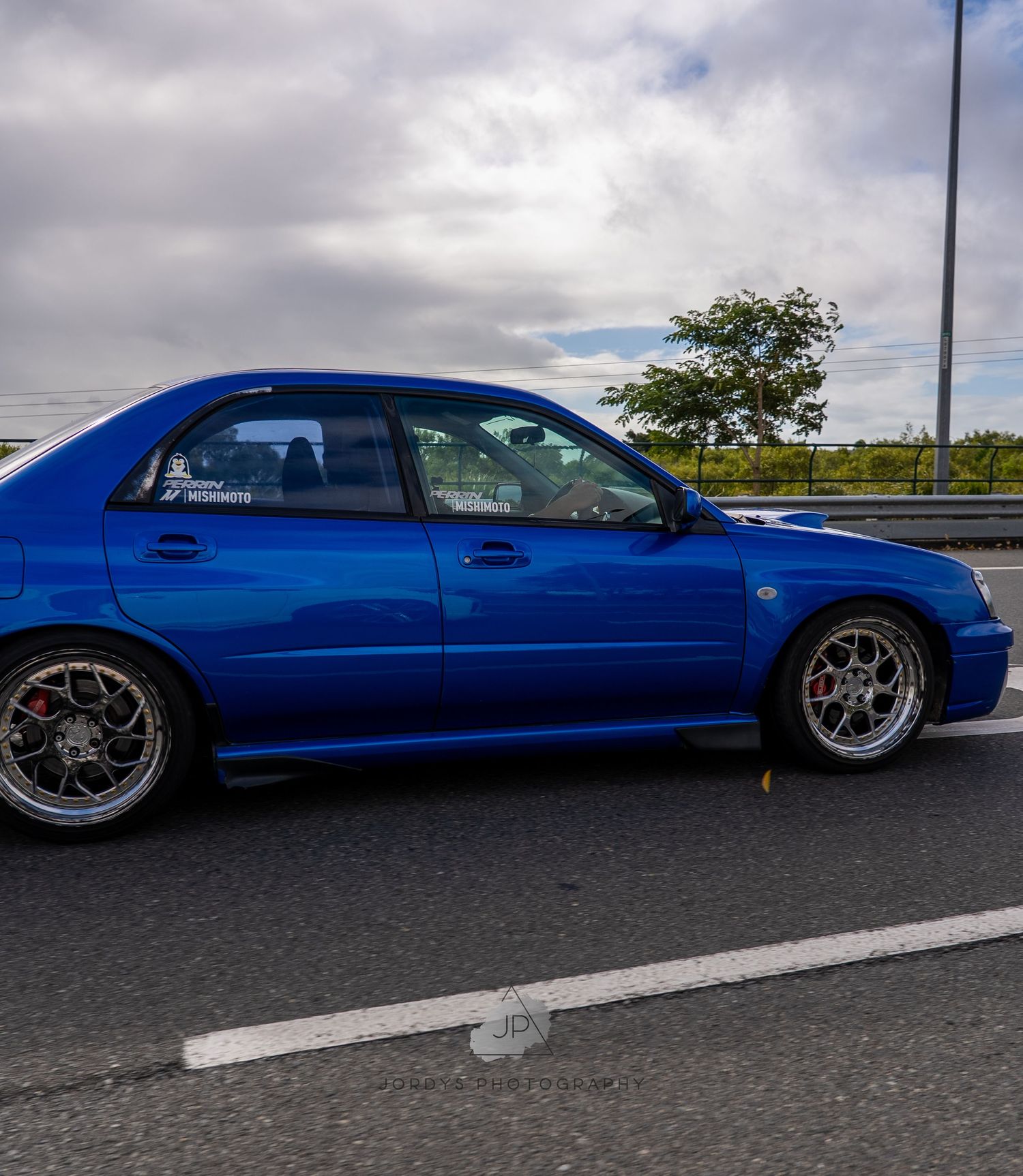 Modified blue Lexus IS300 sedan showcases custom wheels and lowered suspension on a cloudy day in a parking lot.