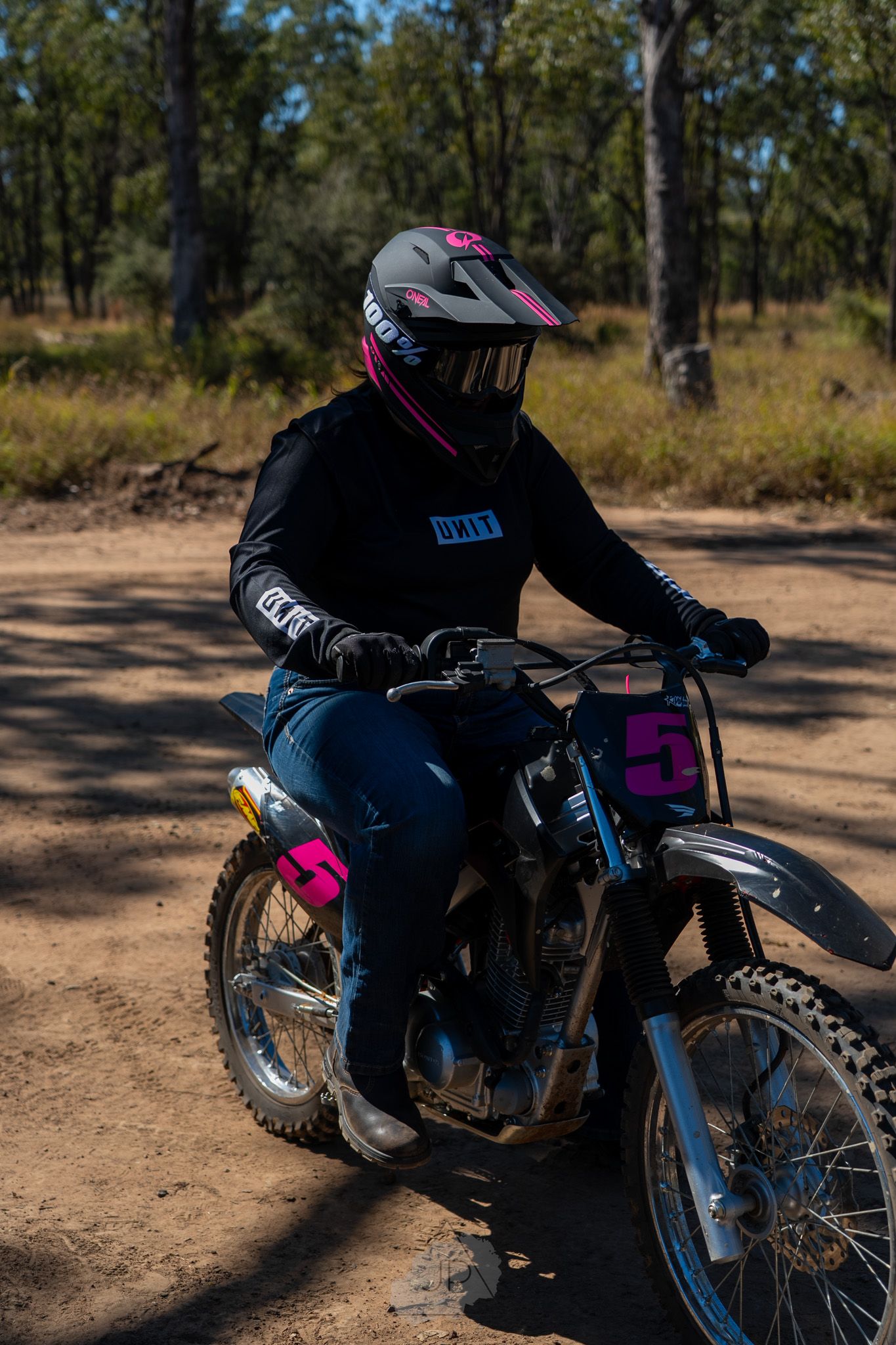 Motocross rider tackles a dirt track through wooded terrain on a black dirt bike with pink accents.
