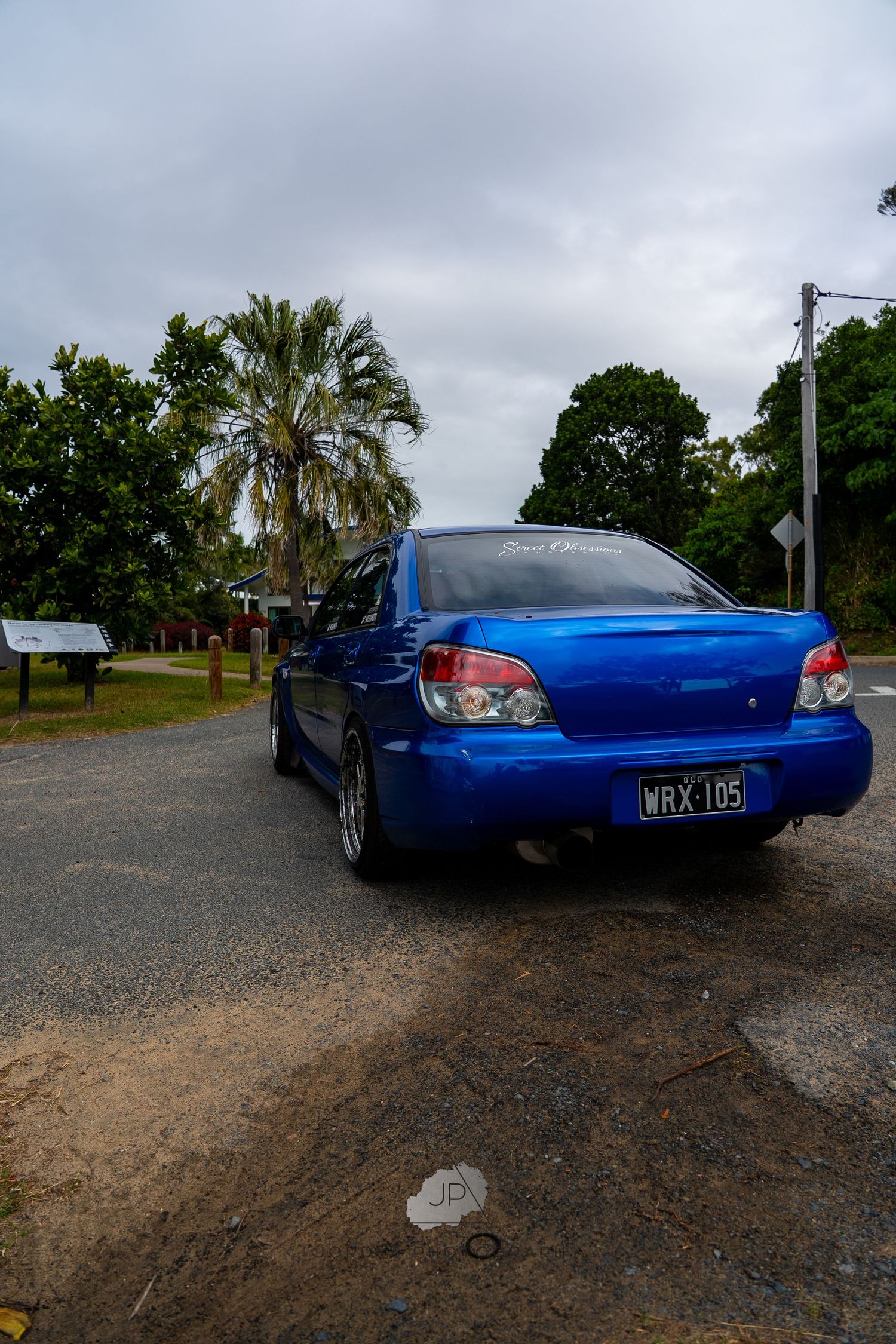 A blue Subaru WRX parked on a dirt road with palm trees and greenery in the background under cloudy skies.