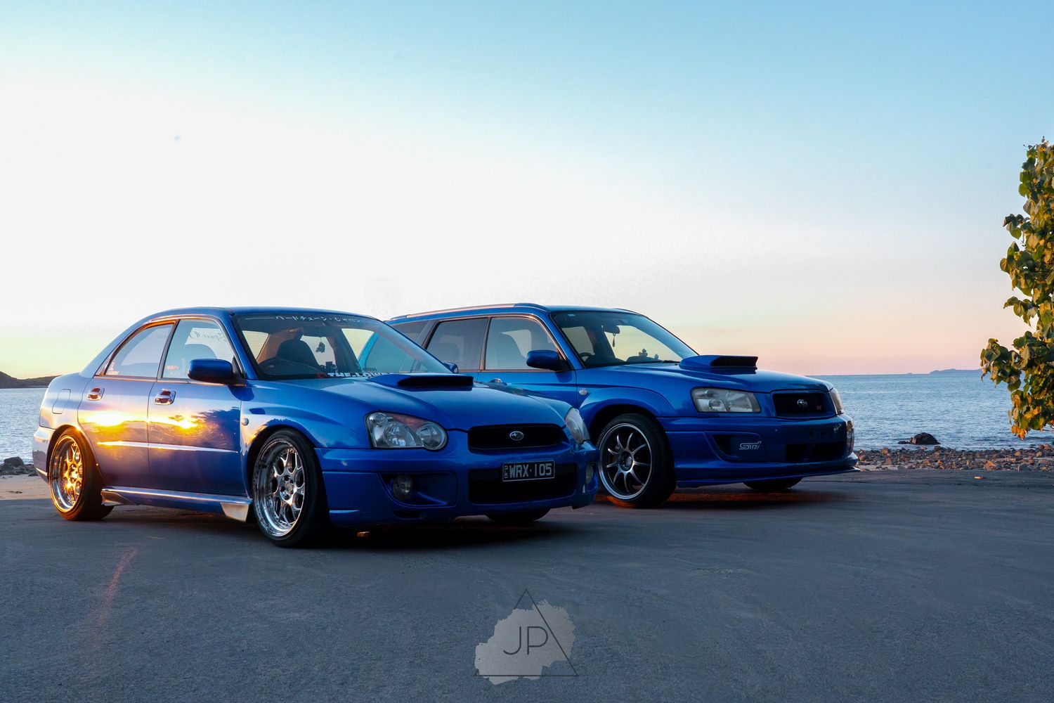 Two blue performance cars parked side by side against a sunset sky with water in the background.
