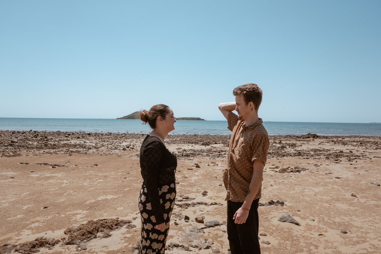 Two people stand on a rocky beach landscape with blue ocean and sky in the background.