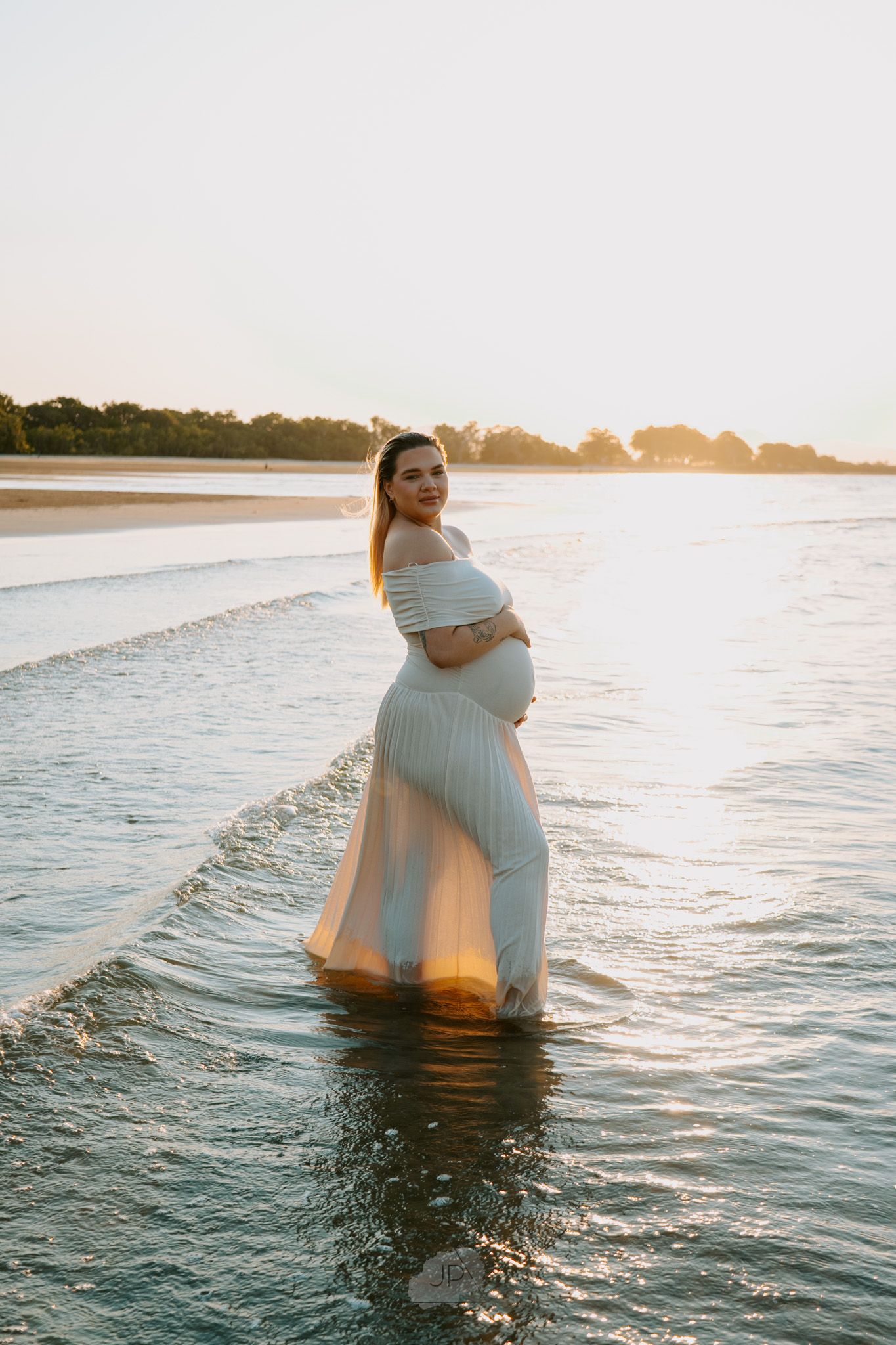 A figure in a flowing white dress stands in shallow ocean water at sunset creating a dreamy silhouette against the horizon.