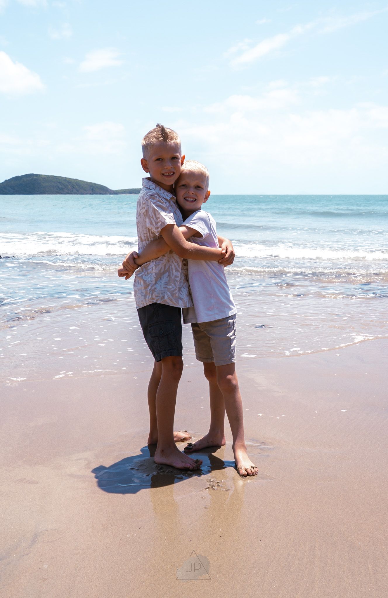 Two children embrace joyfully while playing together on a sandy tropical beach with ocean waves and mountains in background.