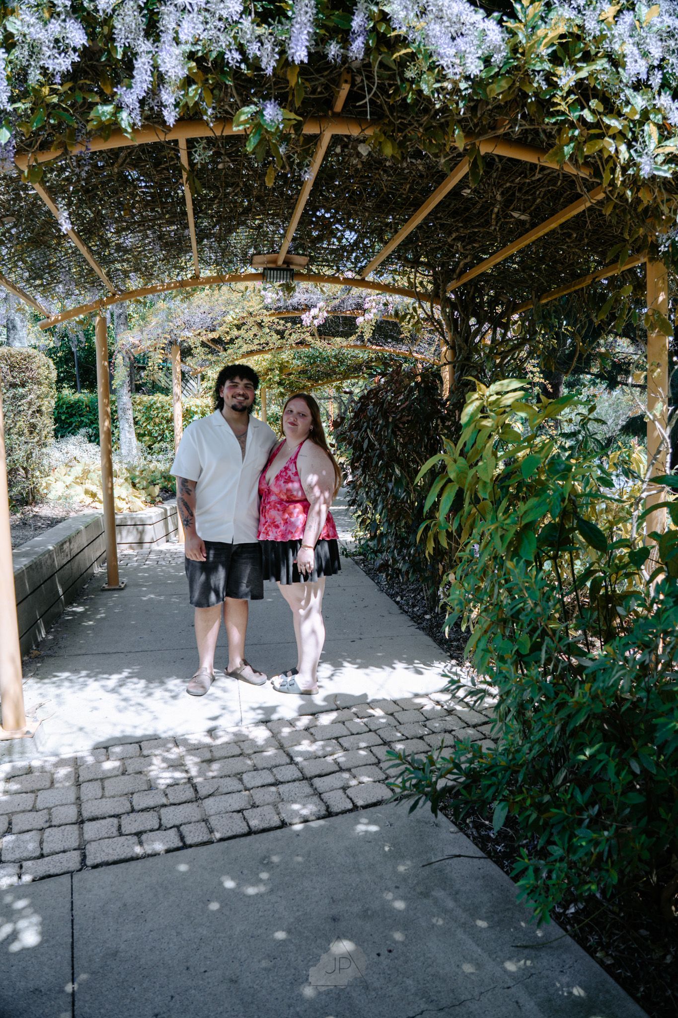 A couple stands under a garden pergola covered in delicate white wisteria flowers on a sunny day.