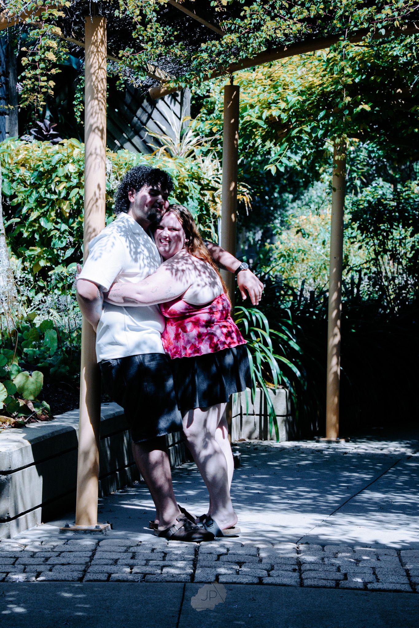 A couple shares a romantic kiss on a stone patio surrounded by lush greenery and wooden posts.