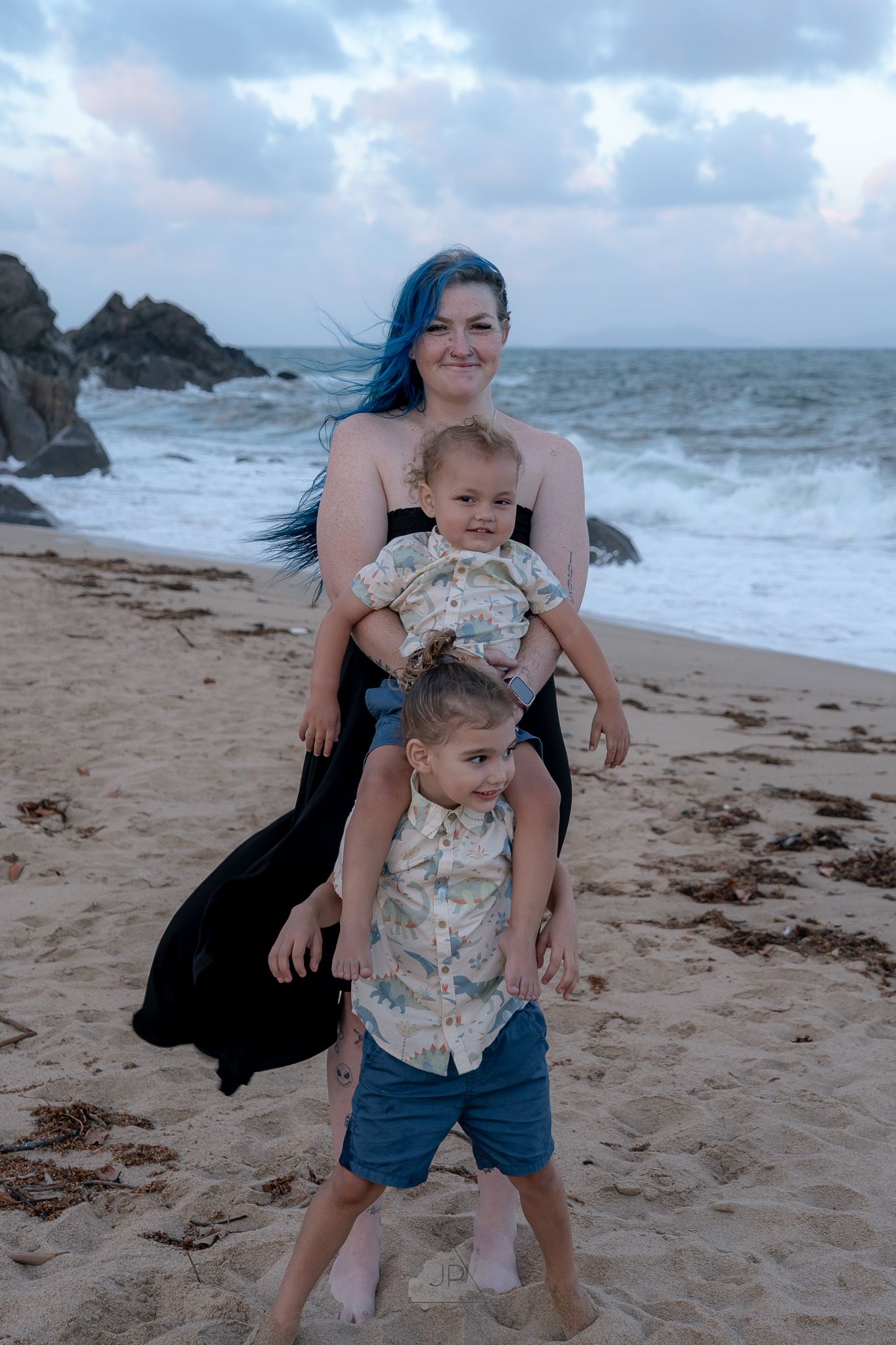 A family enjoys a playful moment together on a sandy beach with crashing waves and rocky cliffs in the background.