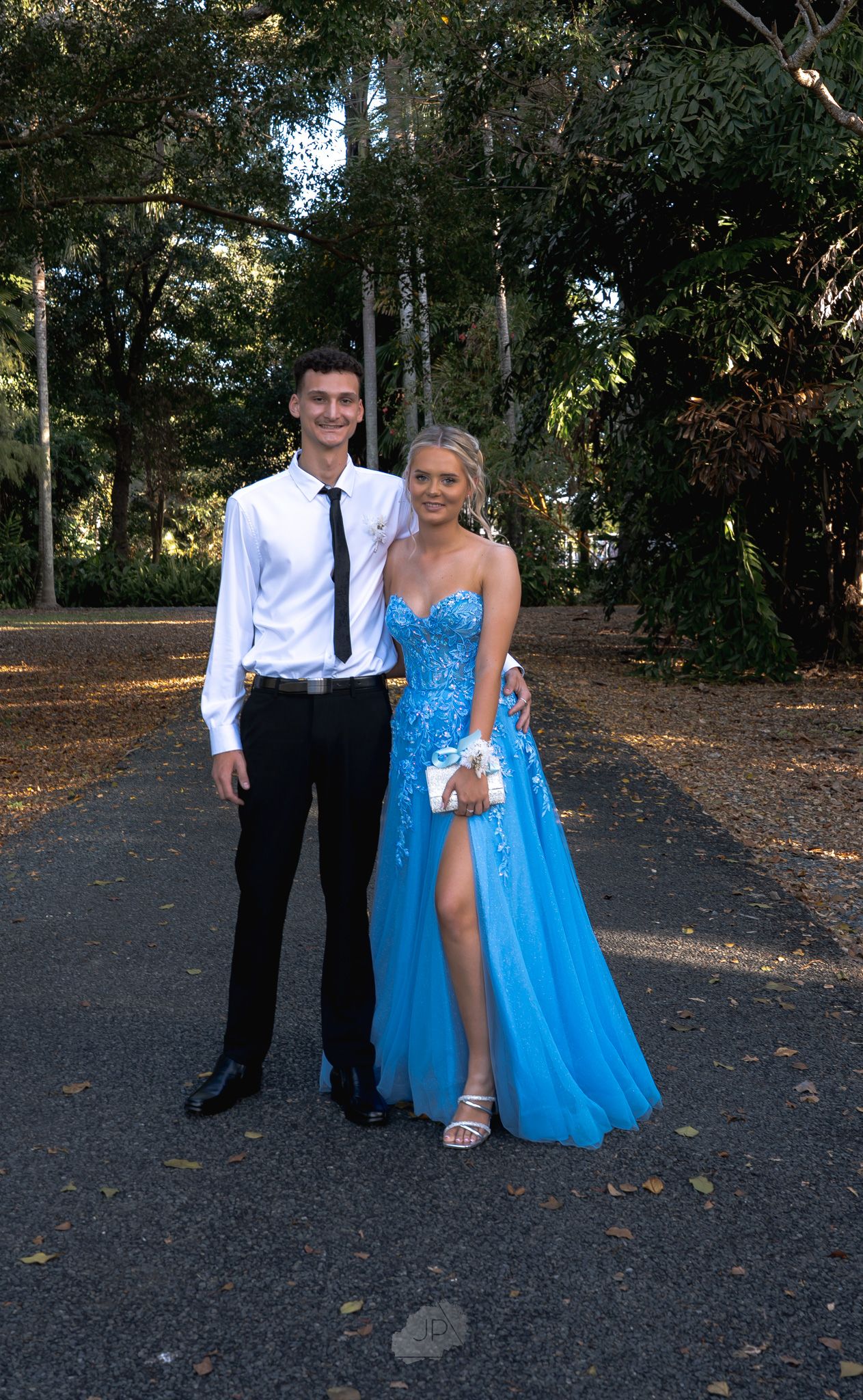 A couple in formal attire poses for photos outdoors, with the person in a bright blue gown and another in a white shirt and tie.