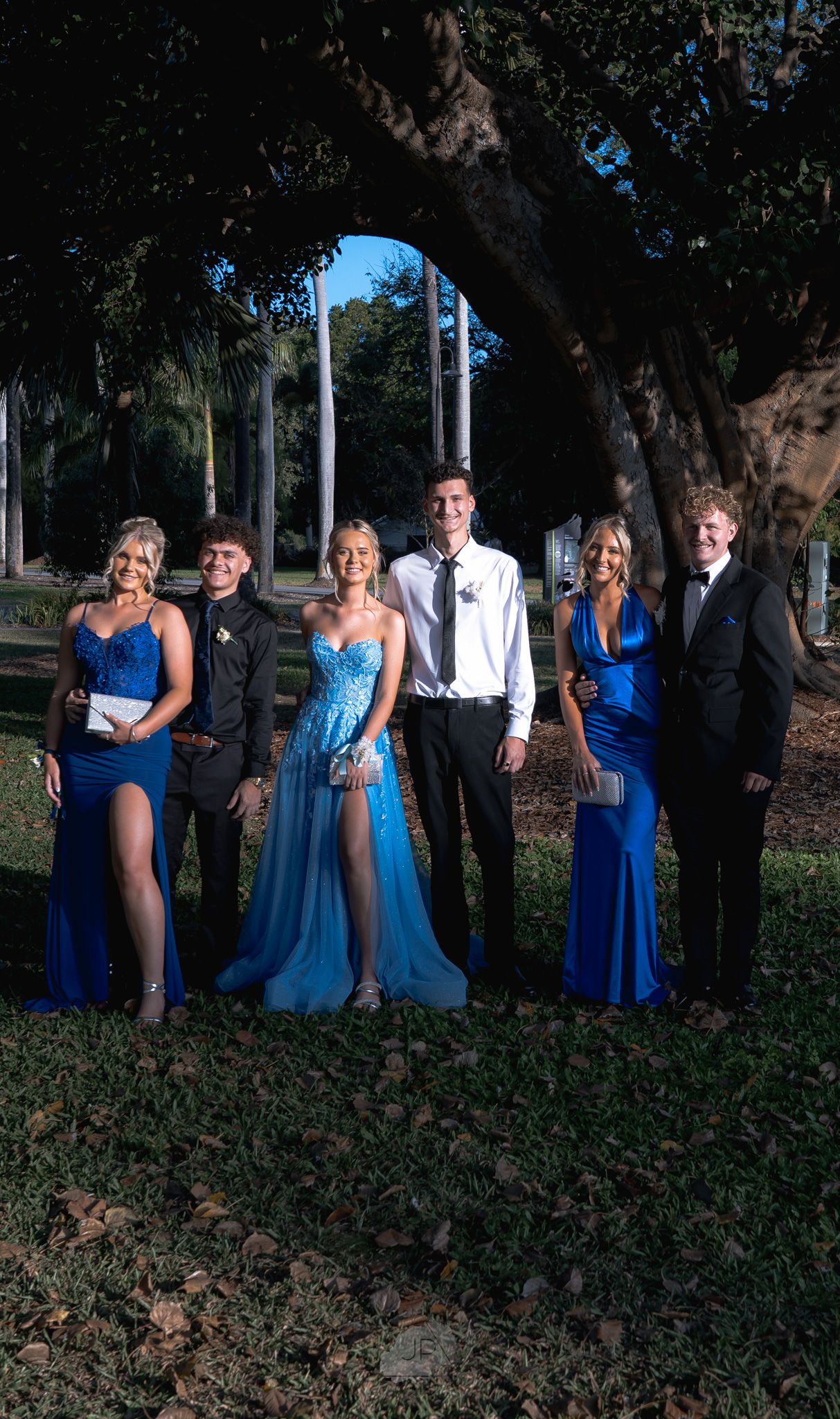 Wedding party members in blue dresses and formal attire pose together in an outdoor garden setting.