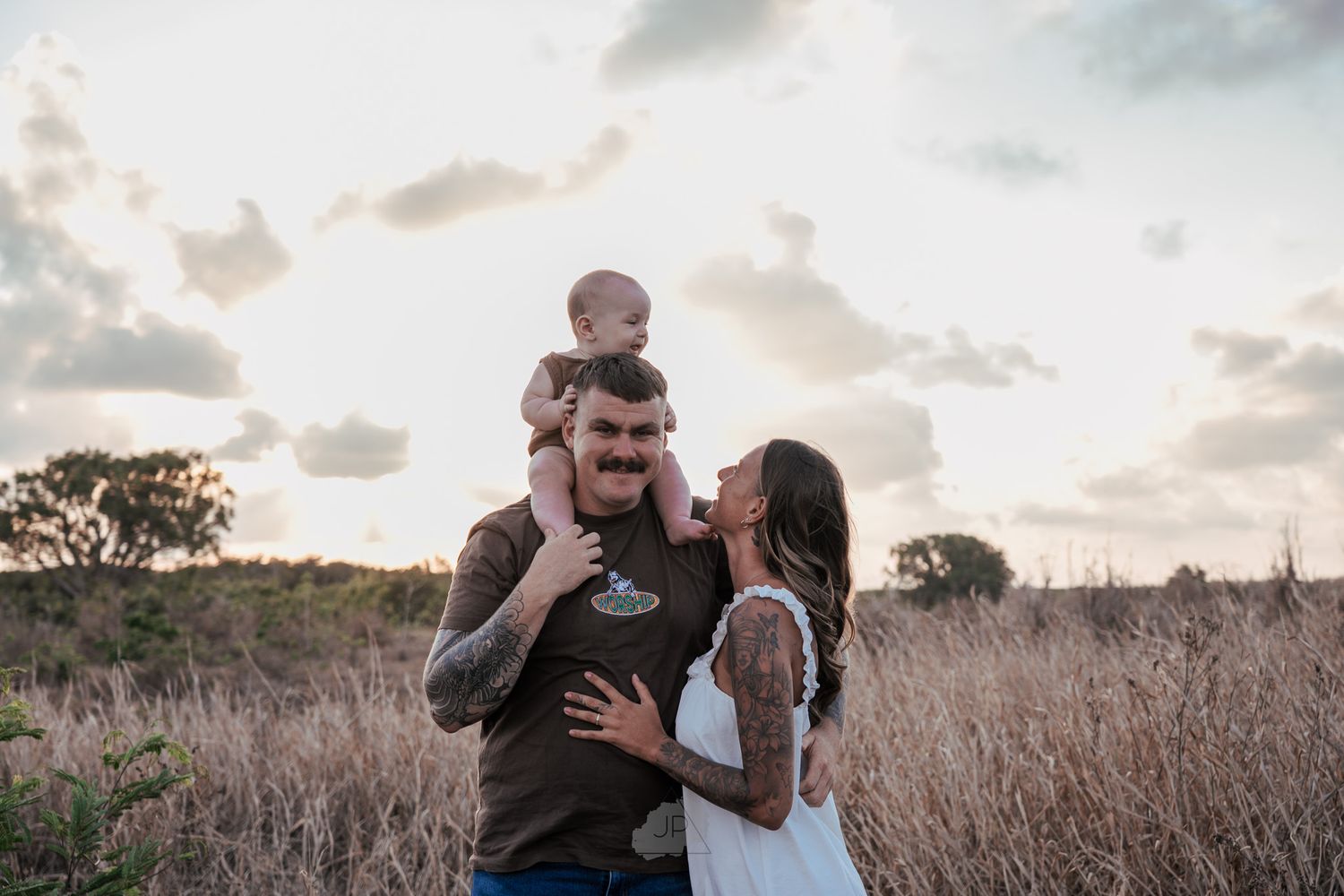 A couple embraces in a golden field during sunset while surrounded by tall grass and trees.