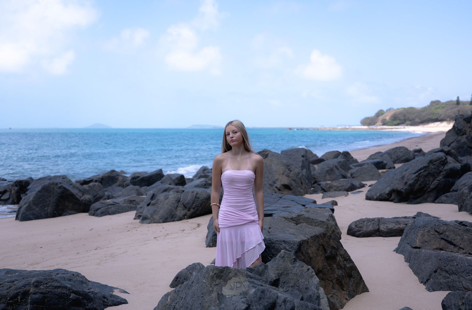 A beachside portrait session featuring someone in a pink dress posing among dark coastal rocks.