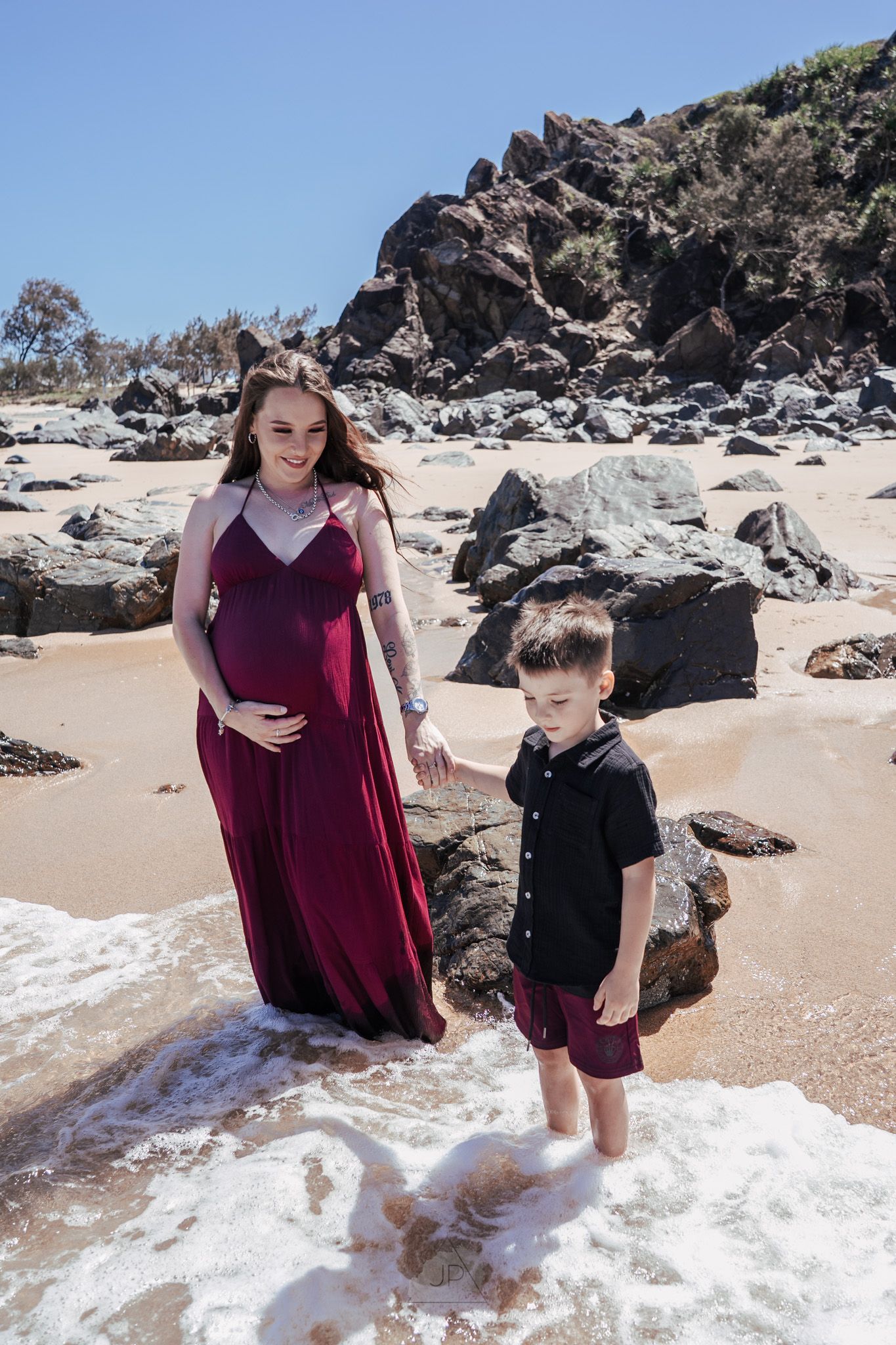 Two people stand on a sandy beach with rocky cliffs and ocean waves at their feet.