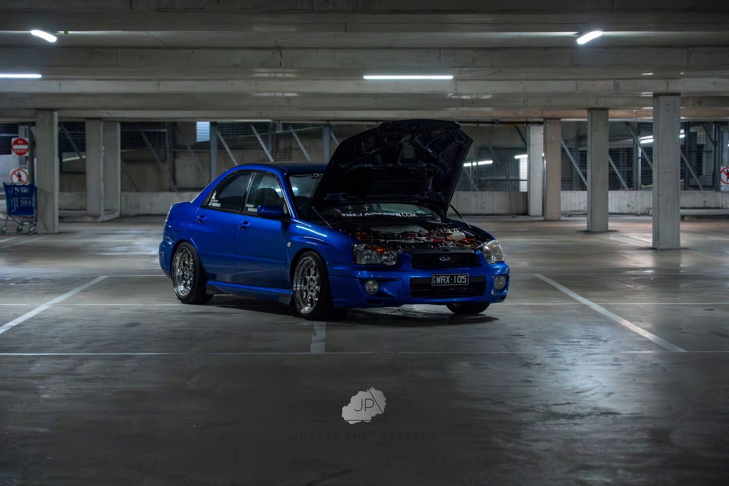 Blue Subaru WRX STI sports car parked in a multi-level concrete parking garage under fluorescent lighting.