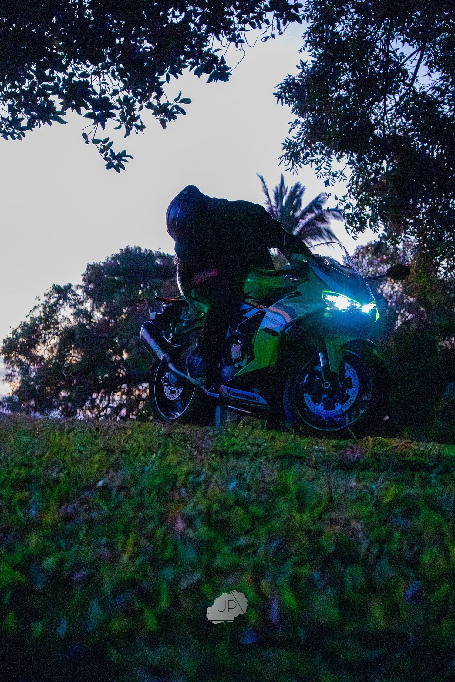 A low-angle silhouette of a green motorcycle set against a sunset-lit sky and tree line at dusk.