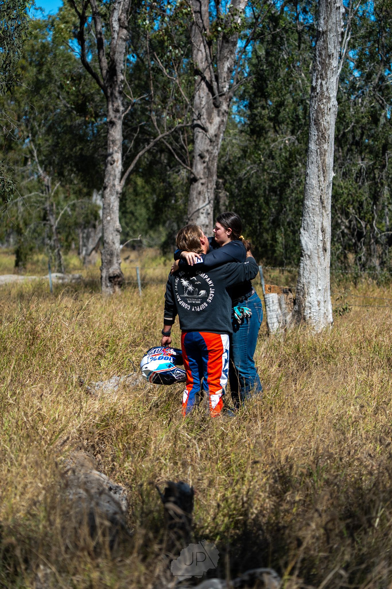 Two people sit close together on grassy ground beneath tall eucalyptus trees on a sunny day.