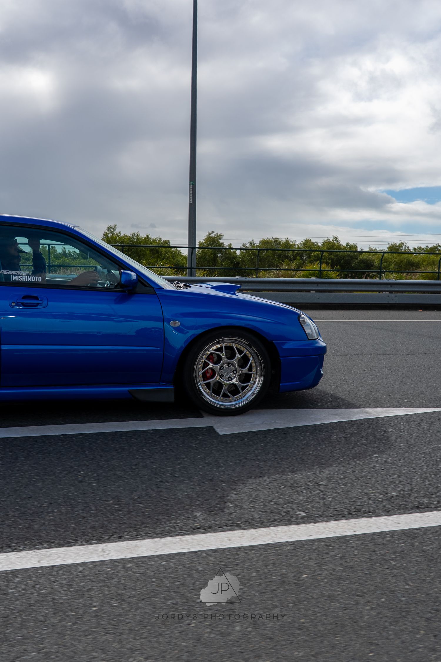 Low angle shot of a modified blue car with custom wheels on a highway against cloudy skies.