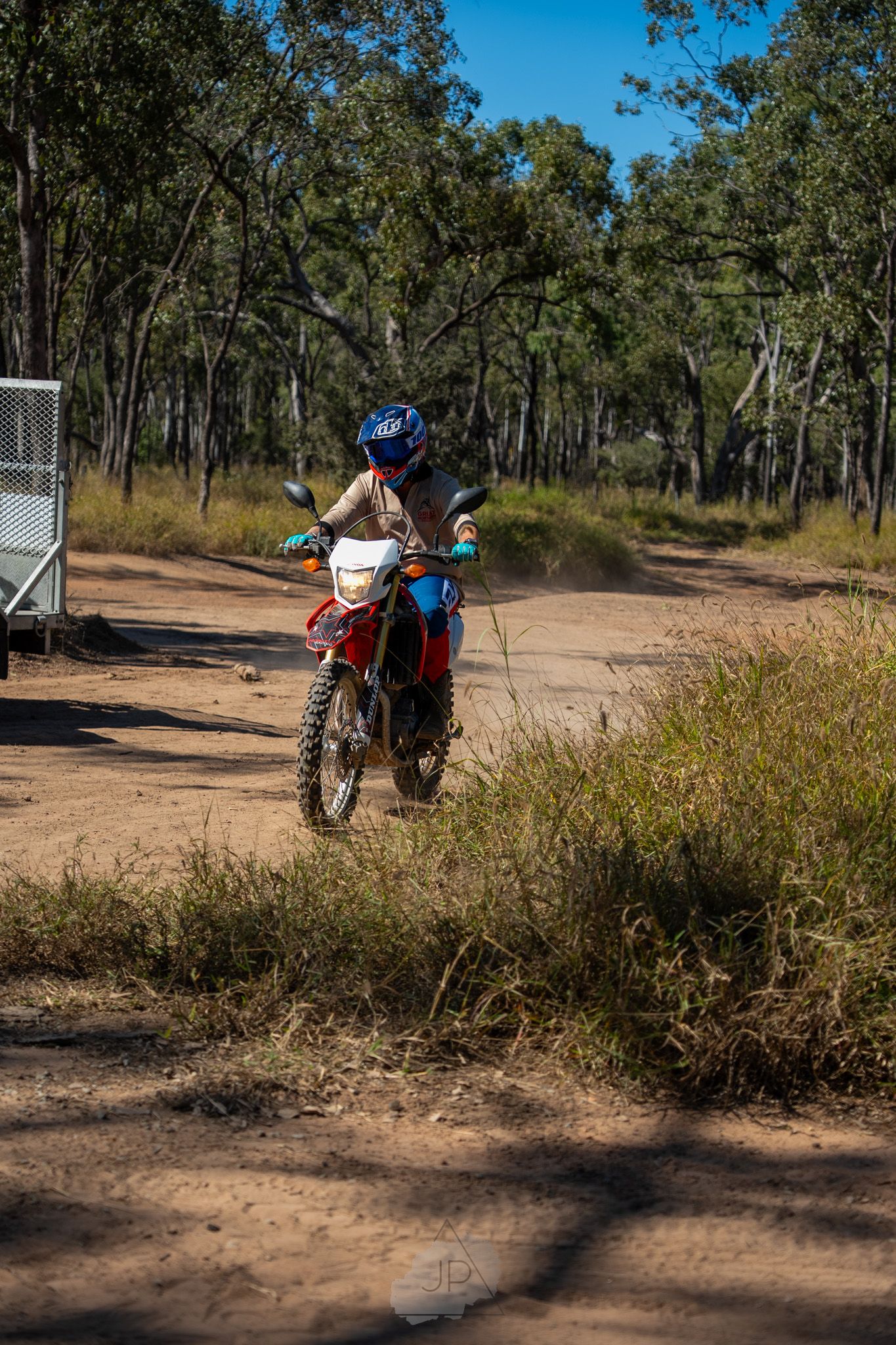 A dirt bike rider performs a dynamic jump on a dusty off-road trail through Australian bushland terrain.
