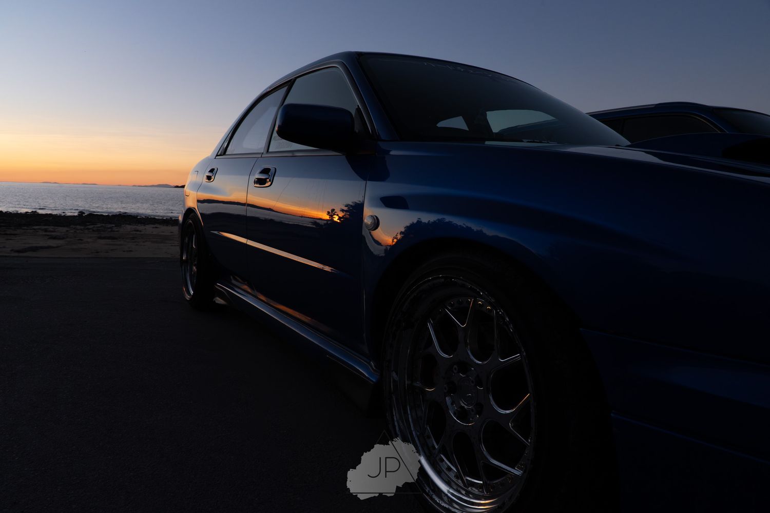 Blue sports sedan parked at sunset with alloy wheels visible against a darkening coastal landscape.