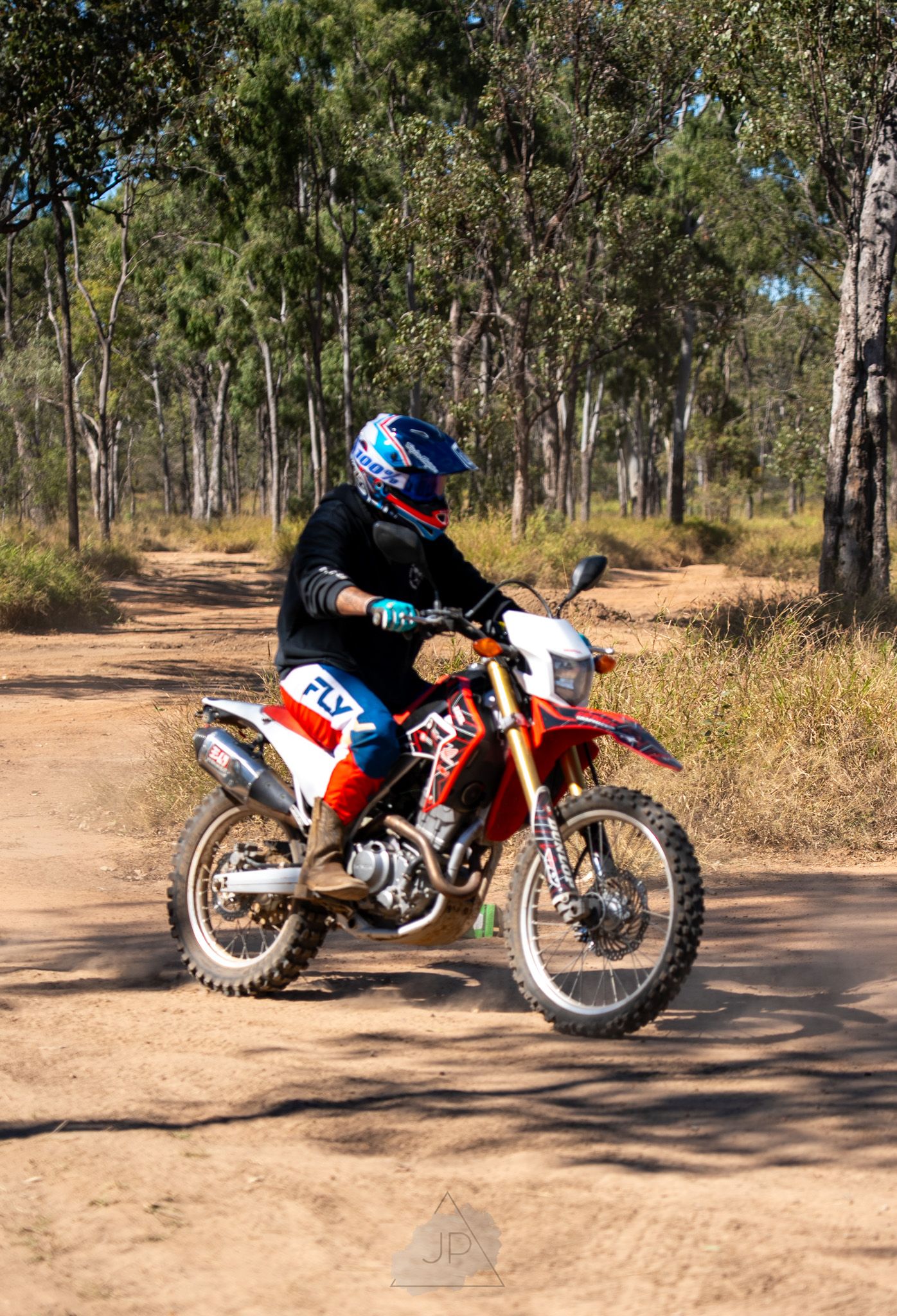 A dirt bike makes a tight turn sequence on a dusty trail through eucalyptus trees in the Australian bush landscape.