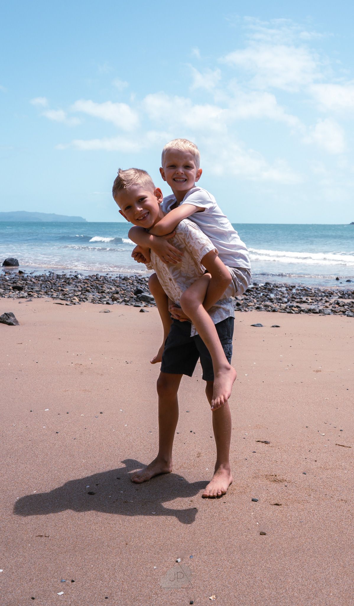 Two young children play and laugh together on a sunny beach with rocky shoreline and ocean waves in the background.