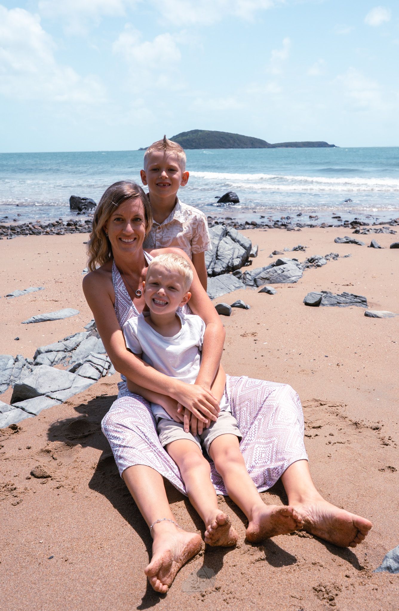 A couple sits together on a sandy beach with waves and rocks in the background on a sunny day.