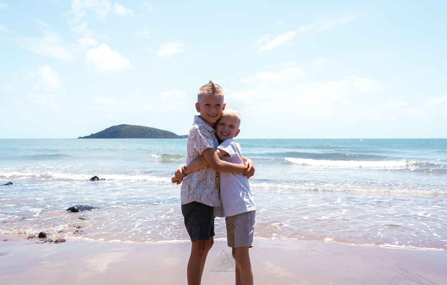 A couple embraces while standing on a sandy beach with ocean waves and blue sky in the background.